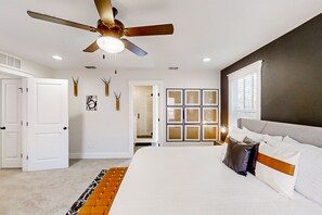 A bedroom with a large bed, a ceiling fan, a window with blinds, and a door leading to another room.