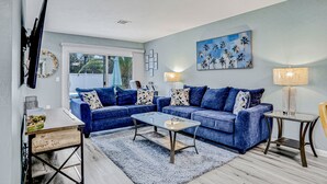 A living room with two blue sofas, a glass coffee table, a side table, and a painting of palm trees.