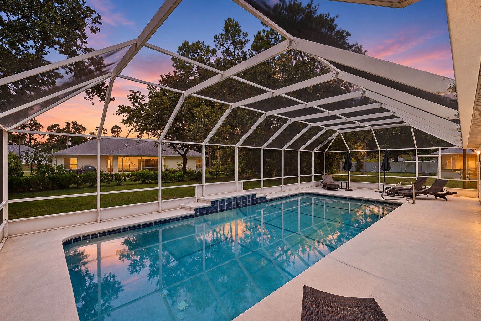 Piscina techada con cerramiento de vidrio, rodeada por una terraza con sillones de descanso y vista a un barrio residencial.