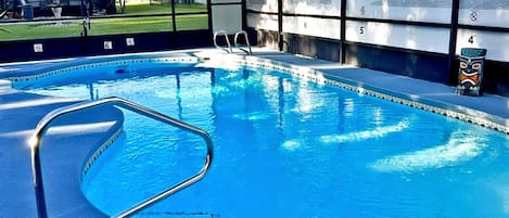 An indoor swimming pool with clear blue water, a metal ladder, and a view of a golf course through large windows.