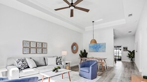 A spacious living room with a white sofa, blue armchair, and a coffee table. A ceiling fan and pendant light are visible. - Brand-new duplex with screened pool, furnished lanai, grill, AC, & washer/dryer (Palm Coast)
