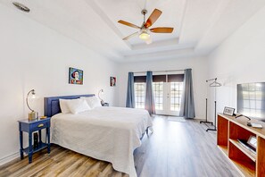 Un dormitorio con cama, ventilador de techo, mesita de noche, televisor y ventana con cortinas.