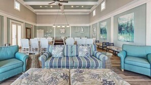 Un salon spacieux avec un grand canapé, deux fauteuils et une table basse. On y trouve un ventilateur de plafond, des luminaires suspendus et des œuvres d’art accrochées aux murs.