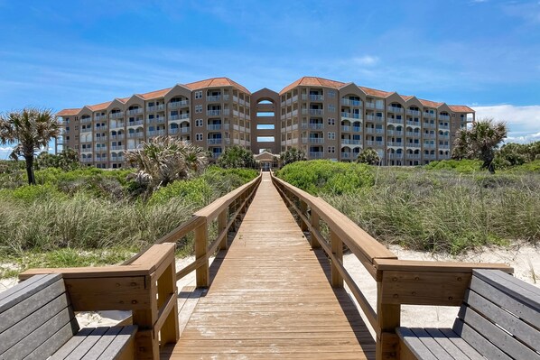 A wooden boardwalk leading to a multi-story hotel with balconies and a central archway.