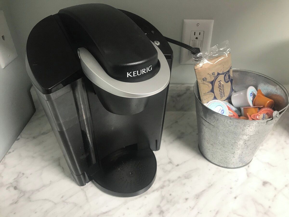 Eine Keurig-Kaffeemaschine auf einer Marmorarbeitsplatte, mit einem Metallbehälter für Kaffeekapseln und einer Kaffeetüte.