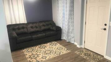 Una sala de estar con un sofá oscuro, alfombras con diseños y una puerta blanca.