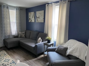 A living room with a sectional sofa, a smaller armchair, a side table with a plant, and a rug. - Cozy One Bedroom House (Adrian)