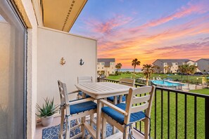 A balcony with a table and chairs, a potted plant, and a view of a swimming pool and buildings during sunset.