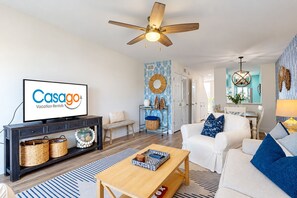A modern living room with a television displaying 'Casago Vacation Rentals,' a ceiling fan, a dining area with a chandelier, and a kitchen visible through an open door.