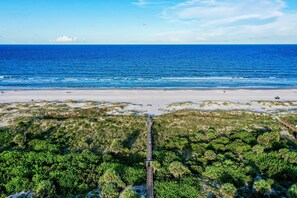 Une plage offrant une vue dégagée sur l’océan, une rive sablonneuse et une promenade en bord de mer qui traverse une zone de végétation côtière.
