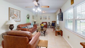 A living room with a brown leather sofa, a coffee table, and a ceiling fan. There are framed pictures on the wall and a TV mounted above a window with blinds. - Spacious Pet -Friendly Home w/ Fenced in Yard and Close to The Beach (St. Augustine)