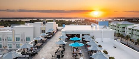 A rooftop terrace with outdoor seating and umbrellas, overlooking a sunset.