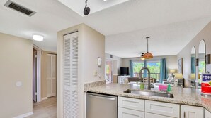 Una cocina con lavavajillas, pileta y encimera. Se ve una ventana con cortinas, un ventilador de techo y una puerta que da a otra habitación.