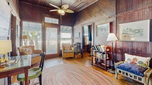 A cozy room with wooden walls, a ceiling fan, a dining table with chairs, a sofa, a bookshelf, and large windows. - Fabulous oceanfront home with ocean view, WiFi, washer/dryer, & central AC (St. Augustine)