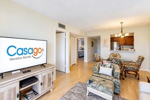 A living room with a television, a dining area with chairs, and a kitchen with wooden cabinets.