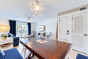 A modern living room with a wooden dining table, blue sofas, a ceiling fan, and a TV mounted on the wall. - A1A South St Augustine | 2 bedroom Oceanview Condo | Sleeps 8 (St. Augustine)