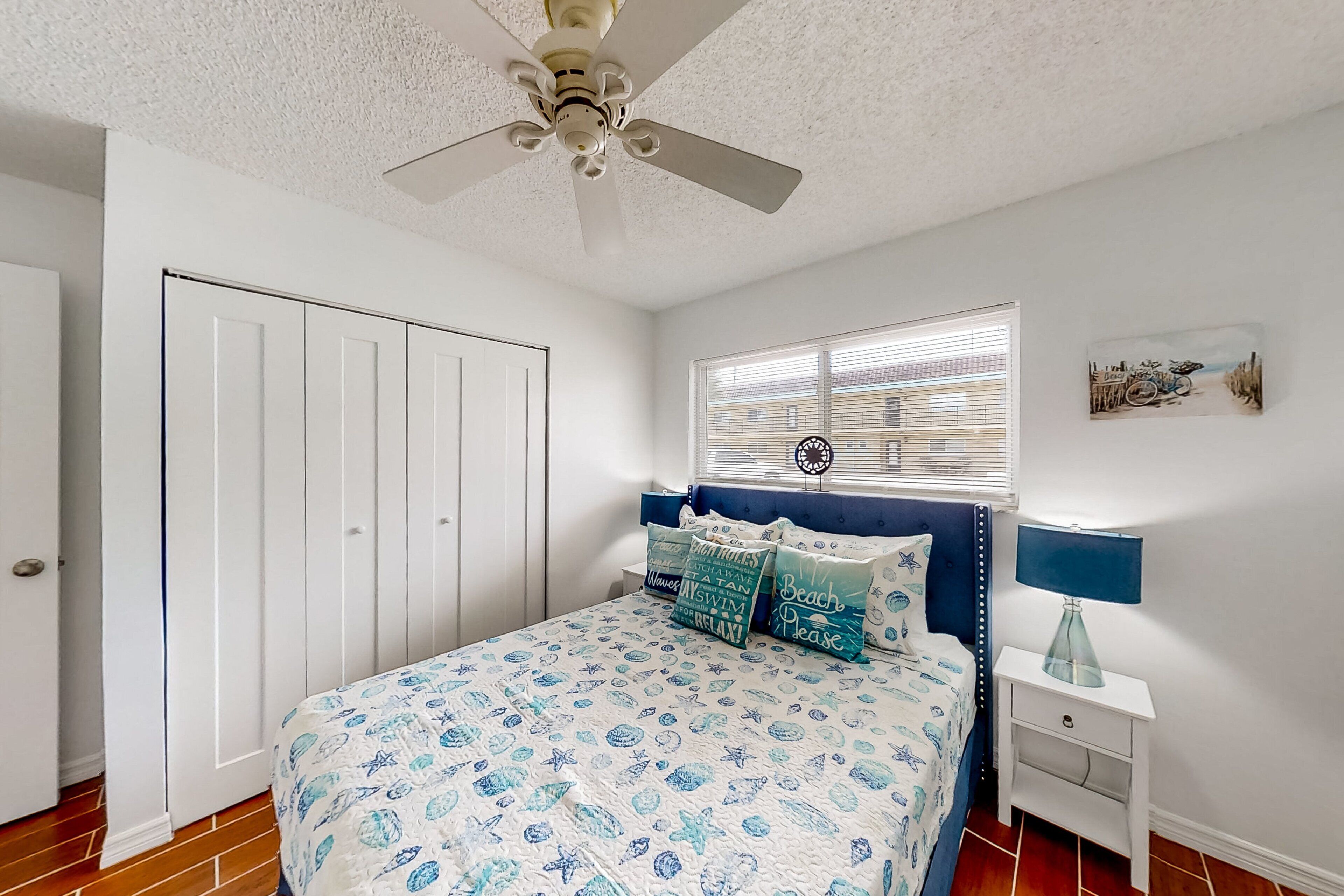 Un dormitorio con cama, mesita de noche, ventilador de techo, ventana con persianas y puerta.