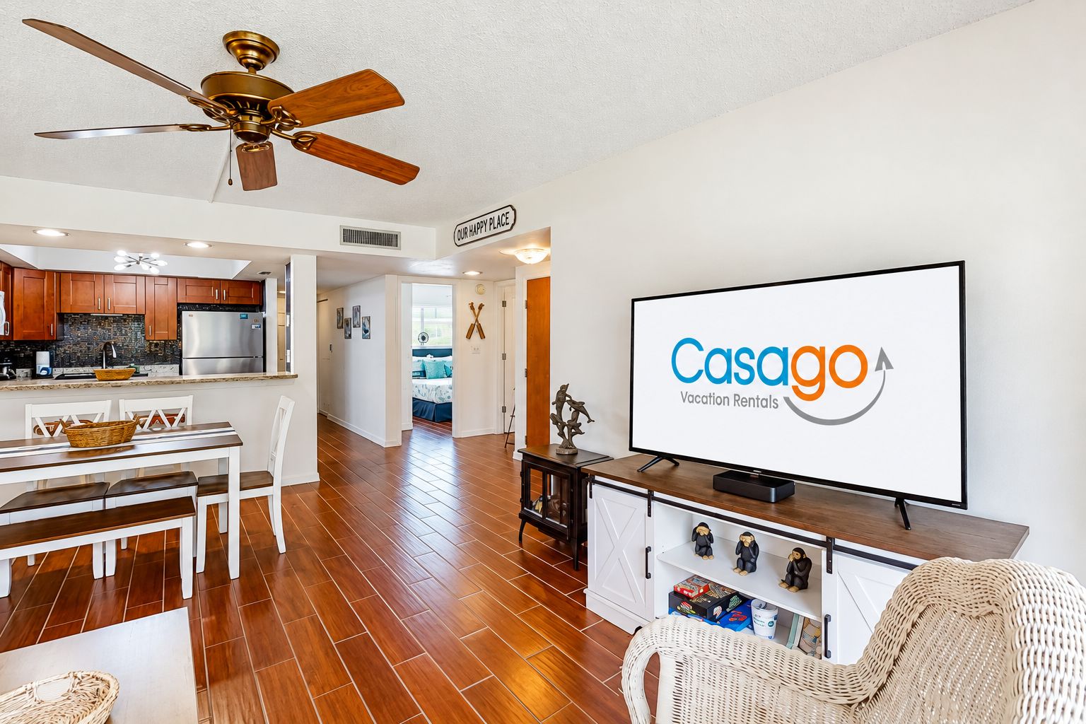 A modern living room with a television displaying 'Casaggo Vacation Rentals,' a dining area with wooden flooring, a kitchen with dark cabinets, and a ceiling fan.