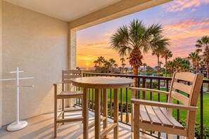 A balcony with a table and chairs, overlooking a sunset view with palm trees and a pool.