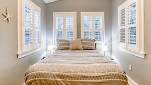 Une chambre avec un grand lit, deux fenêtres avec volets, une décoration en forme d’étoile de mer et une lampe sur la table de chevet.