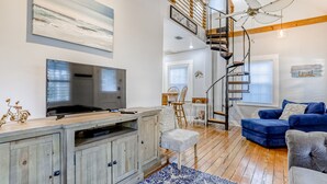 Un salon moderne avec un téléviseur, des meubles en bois, un tapis à motifs bleus et blancs, un canapé bleu et un escalier avec une rampe en bois.