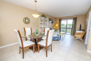 A dining area with a glass table and wooden chairs, a white shelf with decorative items, and a view of a balcony with a railing and a lamp.