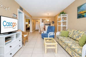 A living room with a television displaying 'Casago Vacation Rentals,' a blue armchair, a patterned sofa, and a white shelving unit. - Lovely Oceanside Home w/Outdoor Shower/Free WiFi/Shared Pool/Hot Tub/Tennis/AC (St. Augustine)