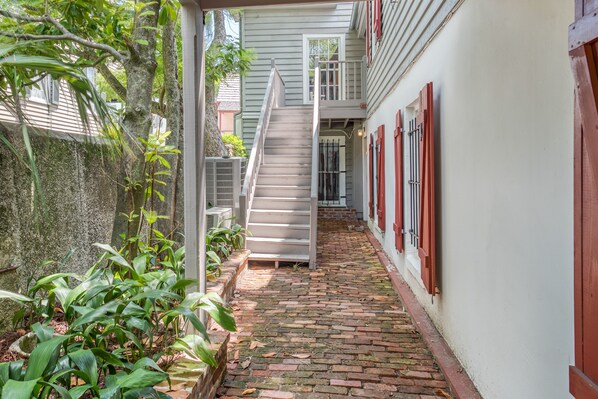 Un étroit sentier pavé de briques mène à un escalier dans une cour dont les murs sont blancs et les volets rouges.