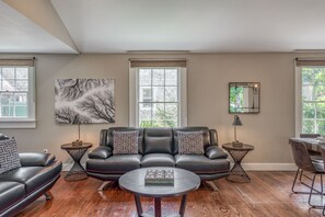 A modern living room with a leather sofa, two side tables, a coffee table, a painting, a mirror, and large windows.