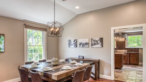 Un coin repas avec une table en bois dressée pour six personnes, entourée de chaises, agrémenté d’un luminaire suspendu et de tableaux encadrés accrochés au mur.