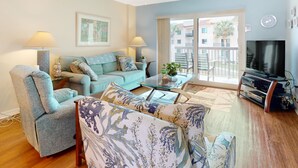 A living room with a sofa, armchair, and coffee table, a TV, and a balcony with a view of buildings.