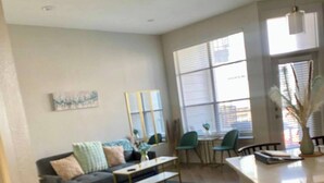 Un amplio salón con sofá, mesa de centro y mesa de comedor. Grandes ventanales ofrecen luz natural y vistas a la ciudad.