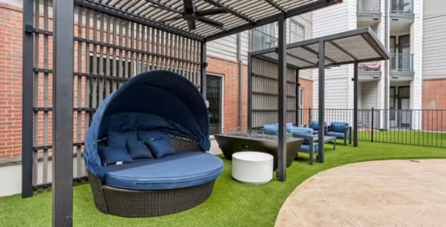 Un espace extérieur aménagé, abrité et doté de sièges, avec un fauteuil suspendu et une table, le tout entouré d’un mur de briques et d’une clôture métallique.
