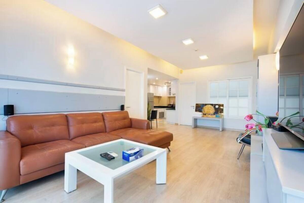 A modern living room with a leather sofa, a glass coffee table, and a kitchen in the background.