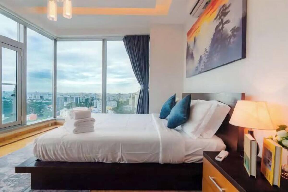 A modern hotel room with a large bed, a nightstand, a lamp, and a city view through large windows.