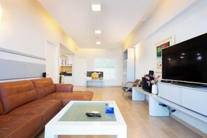 A modern living room with a brown leather sofa, a white coffee table, a flat-screen TV, and a kitchen area in the background.