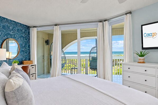 A bedroom with a large bed, a dresser, a mirror, and a view of the ocean through a sliding glass door.