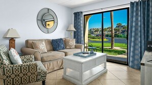 Una sala de estar con un sofá color beige, un sillón estampado y una mesa de centro blanca. Se ve un ventanal con vistas a una carretera y áreas verdes.