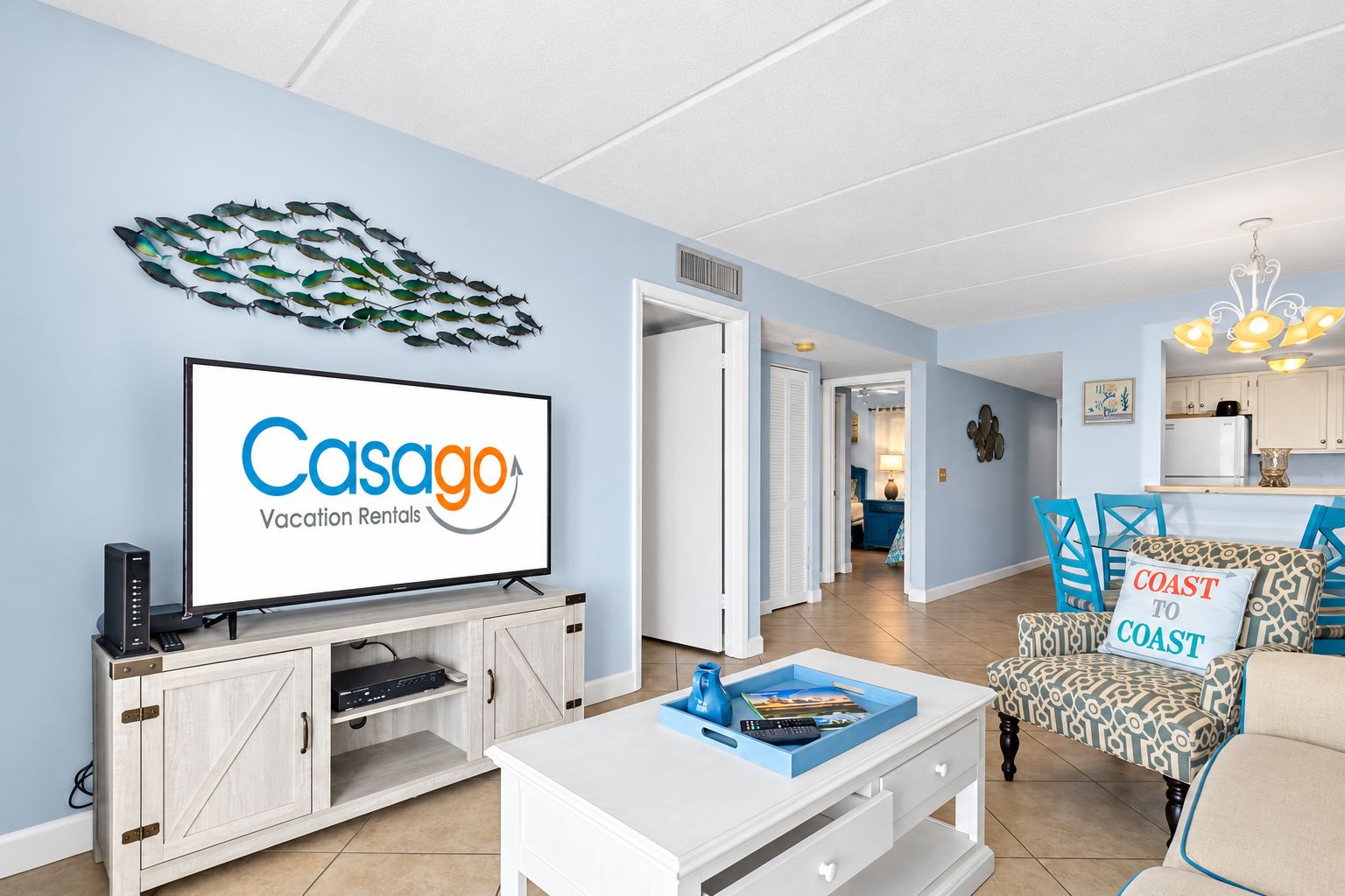 A living room with a television displaying 'Casago Vacation Rentals,' a dining area with blue chairs, and a kitchen in the background.