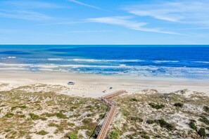 Una playa con un paseo marítimo de madera que lleva al agua, dunas de arena y un cielo azul despejado.