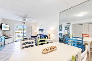 A modern living room with a sofa, dining table, and chairs, a television, and a ceiling fan. - Sunny ground-floor condo w/private patio, W/D, on-site pools, tennis court, BBQ (St. Augustine)