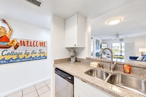 A modern kitchen with granite countertops, stainless steel sink, and a dishwasher. A welcome sign is visible on the wall. - Sunny ground-floor condo w/private patio, W/D, on-site pools, tennis court, BBQ (St. Augustine)