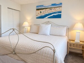 A bedroom with a bed, two bedside lamps, a painting of a beach scene, and a door with louvered shutters. - Sunny ground-floor condo w/private patio, W/D, on-site pools, tennis court, BBQ (St. Augustine)