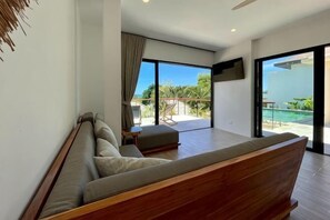 A modern bedroom with a large bed, a small table, and a view of a pool and greenery.