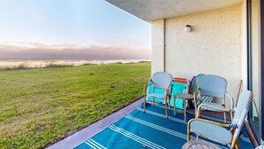 A balcony with wicker chairs and a table, overlooking a grassy area and a distant shoreline. - First floor oceanfront condo with beach access and tennis courts (St. Augustine)