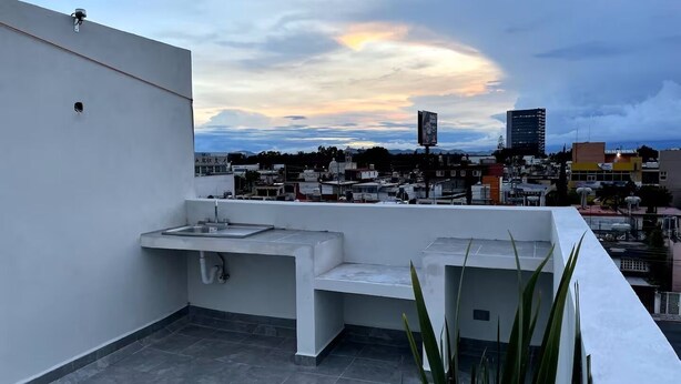 A rooftop terrace with a sink and a small seating area, overlooking a cityscape with buildings and a cloudy sky. - Living Universidad (Puebla)
