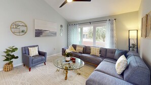 A living room with a sofa, armchair, glass coffee table, ceiling fan, and a large window with curtains. - Spacious condo with 2 private balconies, beach access and pool (St. Augustine)
