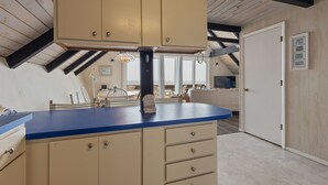 Eine Küche mit blauer Arbeitsplatte, beigen Schränken und einer schwarzen Mittelachse. Rechts ist eine Tür und im Hintergrund ein Fenster mit Jalousien.