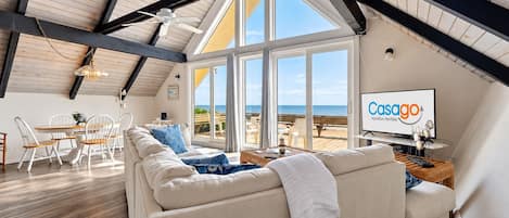 A spacious living room with a large window overlooking the beach, a white sofa, and a television displaying 'Casago Vacation Rentals'.