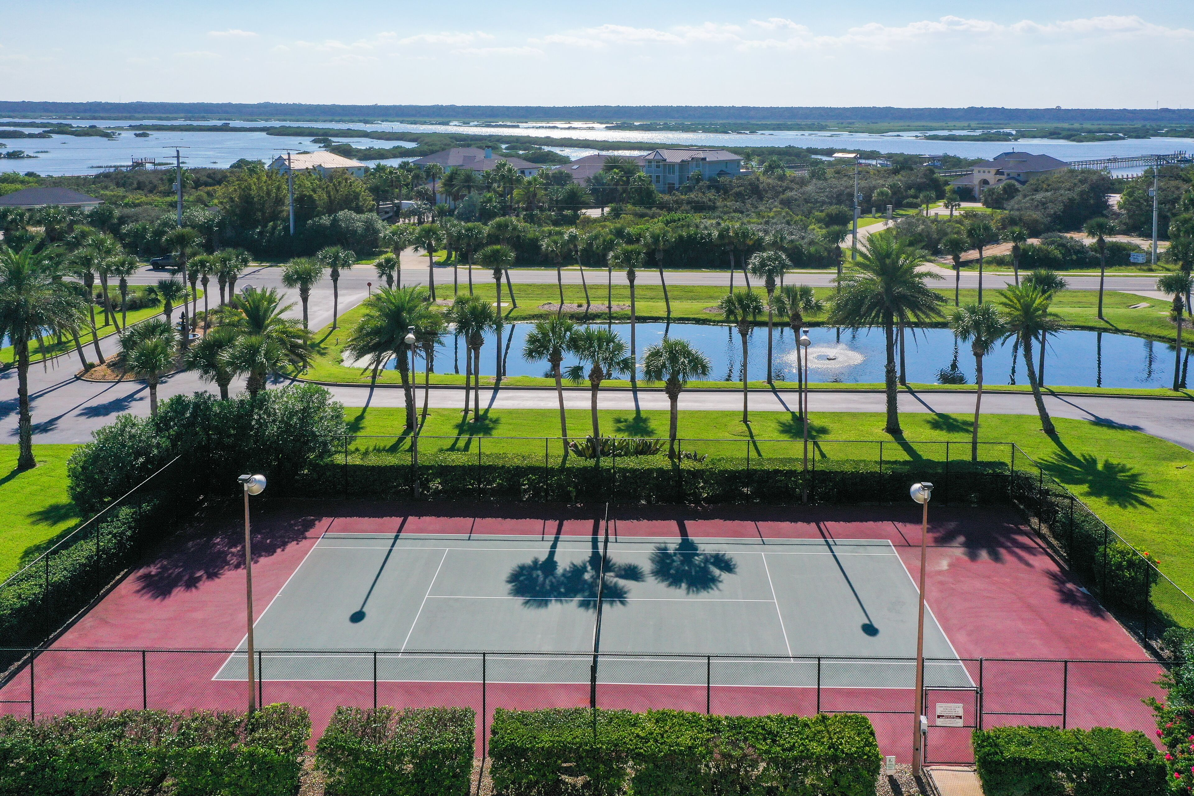 A tennis court surrounded by palm trees and a body of water in the background.